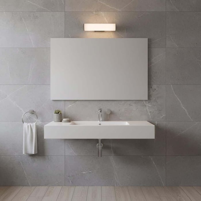 Minimal gray-tiled bathroom lights wall with a wide mirror, small wall light above it, wall-mounted rectangular sink, and a towel ring with a white towel.
