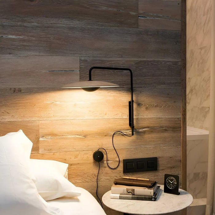 Bedside reading light mounted on a wood-panel wall, casting a warm pool of task light over books and pillows.
