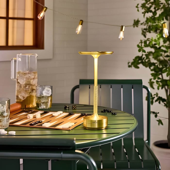 Slim brass outdoor table lamp on a green metal table with backgammon board, ice pitcher, and string lights above.