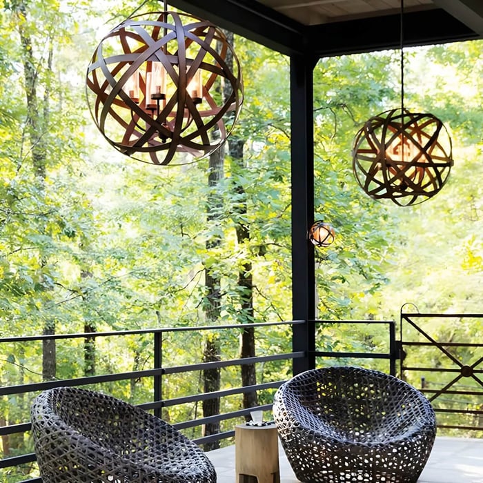 Spherical metal outdoor pendant lights hanging on a covered balcony with forest views and woven chairs.