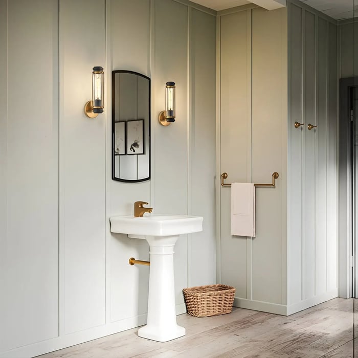 A bathroom space showcasing a pedestal sink and an arched mirror flanked by brass-finished wall sconces. Soft paneling on the walls and minimalistic decor, including a wicker basket and towel rack, create a cozy, inviting atmosphere.