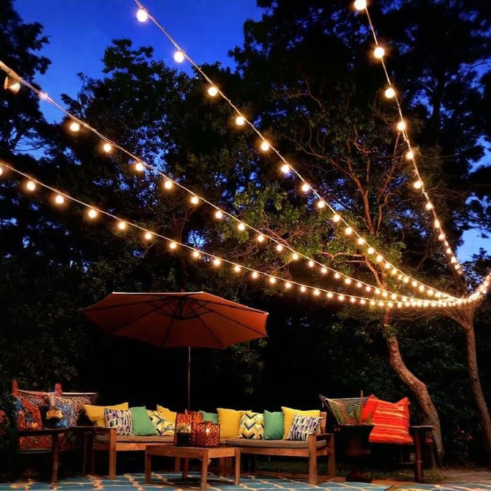 Outdoor string lights draped in zig-zag lines from a tree over a garden lounge area with colourful cushions at night.