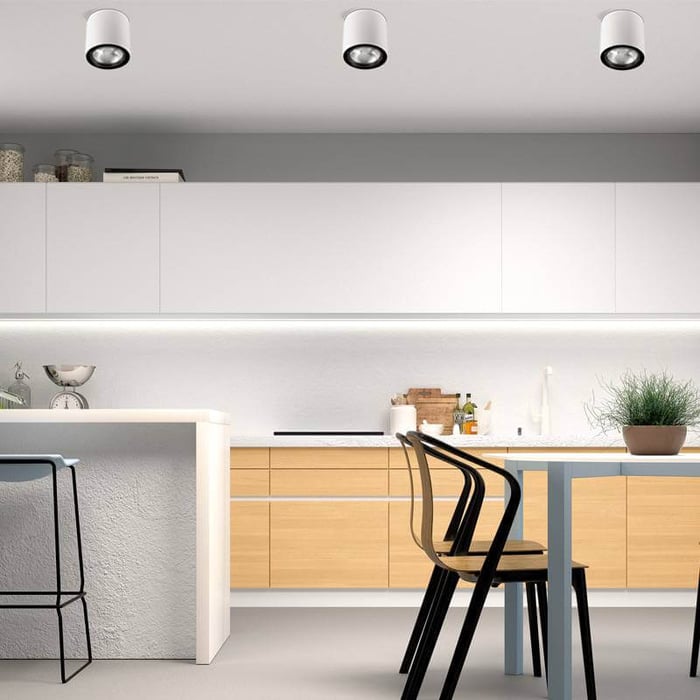 Modern kitchen with a clean, minimalist design featuring white cabinets, a light wood-tone island, and a dining table with black chairs. The ceiling has three cylindrical recessed spotlights, and a strip of under-cabinet lighting provides additional illumination, creating a bright and airy atmosphere. A potted plant and kitchen accessories add a touch of warmth and greenery.