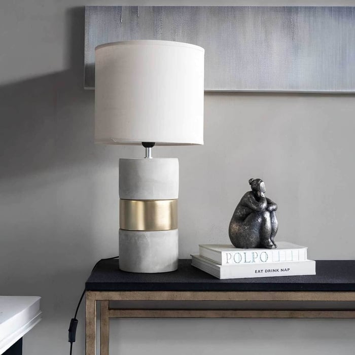Concrete and brass industrial table lamp with a white drum shade on a console table.