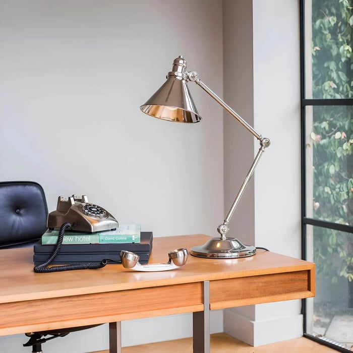 Chrome adjustable swing-arm desk lamp on a wooden table near a window, styled with books and a vintage phone.