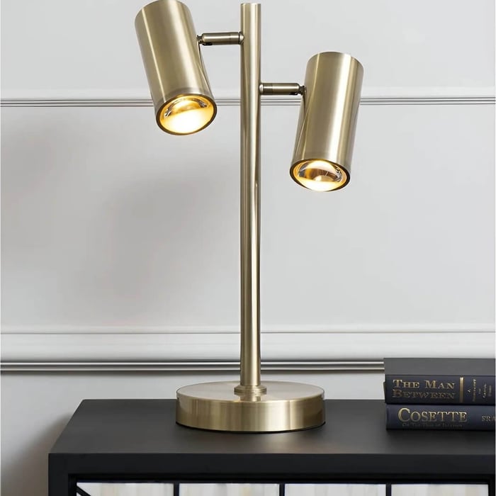 Brass dual-head table lamp on black cabinet with books against a white paneled wall.