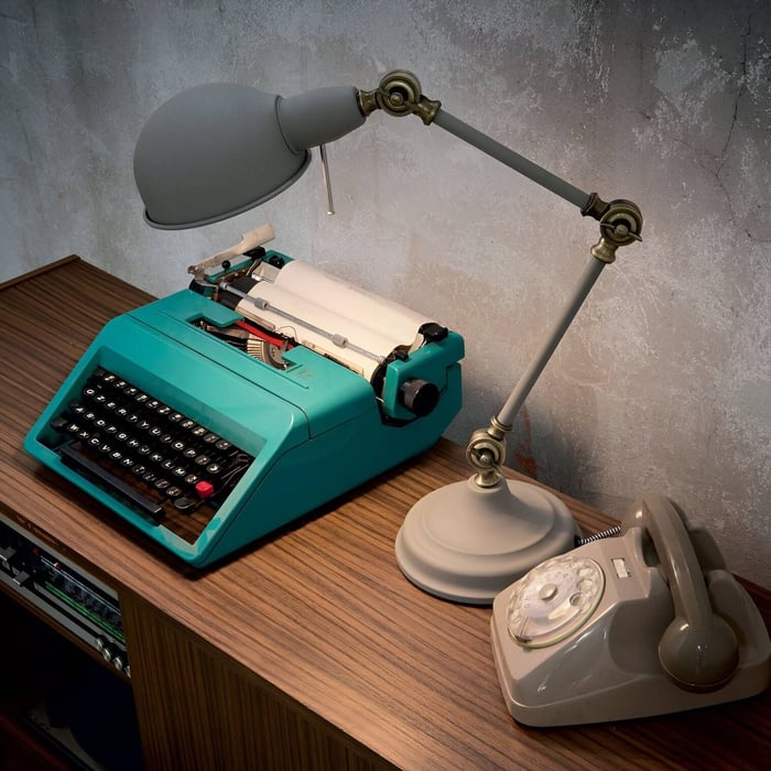 Adjustable grey desk lamp with vintage brass joints positioned beside a turquoise typewriter and rotary phone on a wooden desk.