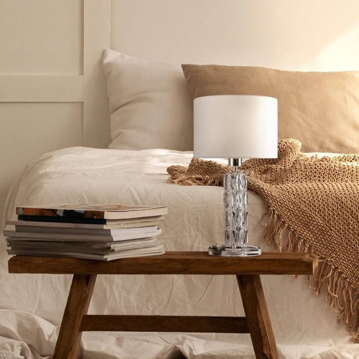Crystal base table lamp with a white shade on a wooden bench at the foot of a bed.