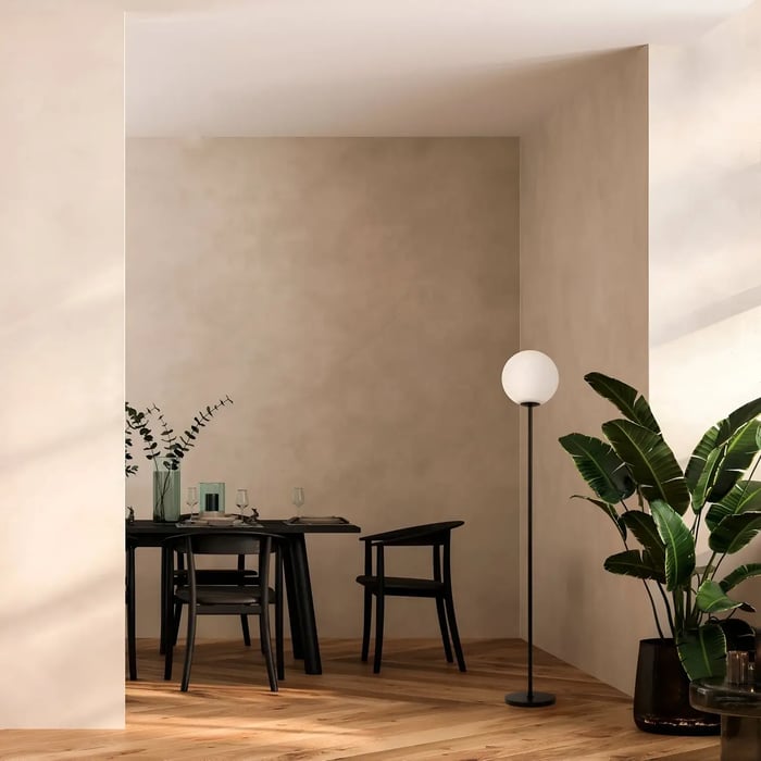 A simple black floor lamp with a spherical white shade illuminating a dining area with a minimalist table and chairs, complemented by greenery and a neutral-toned wall.