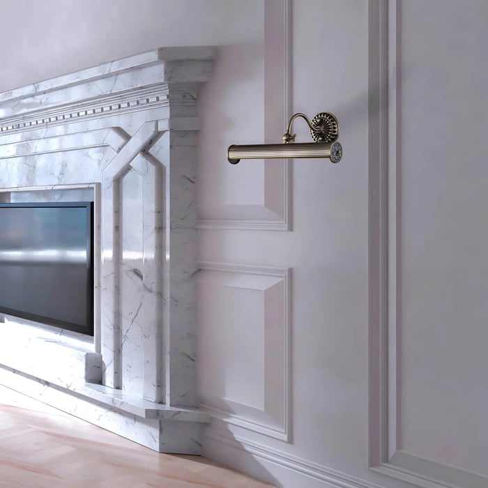 Classic brass wall-mounted picture light extending from a white panelled wall next to a marble fireplace.