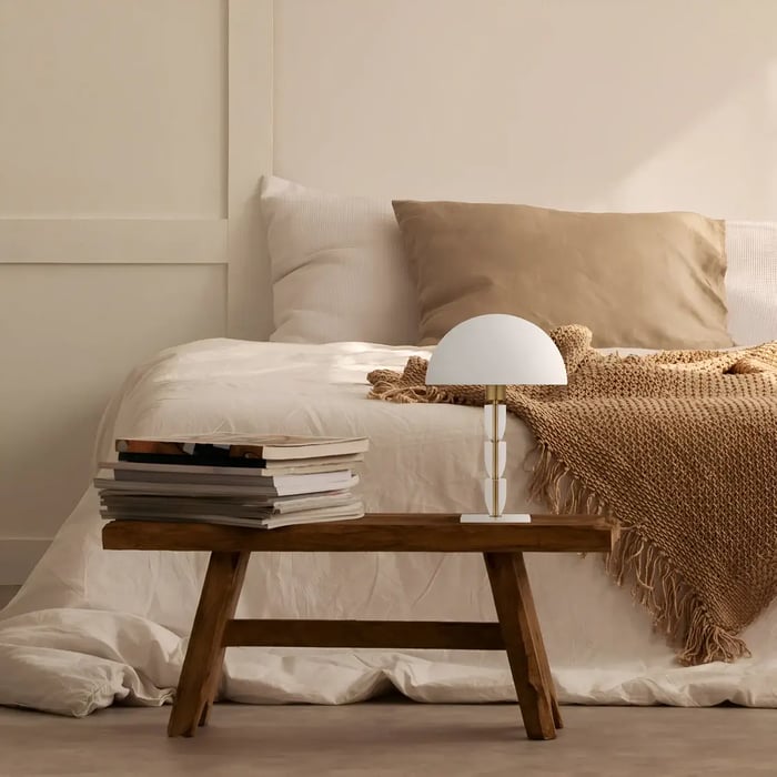 A rustic-inspired bedroom featuring a wooden bench used as a nightstand, topped with a stack of books and a table lamp with a white dome shade, surrounded by beige and neutral bedding and soft textures.