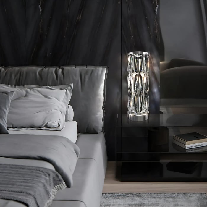 A luxurious bedroom featuring a sleek black bedside table adorned with a crystal table lamp emitting a warm glow, paired with grey bedding and a dark, textured wall.