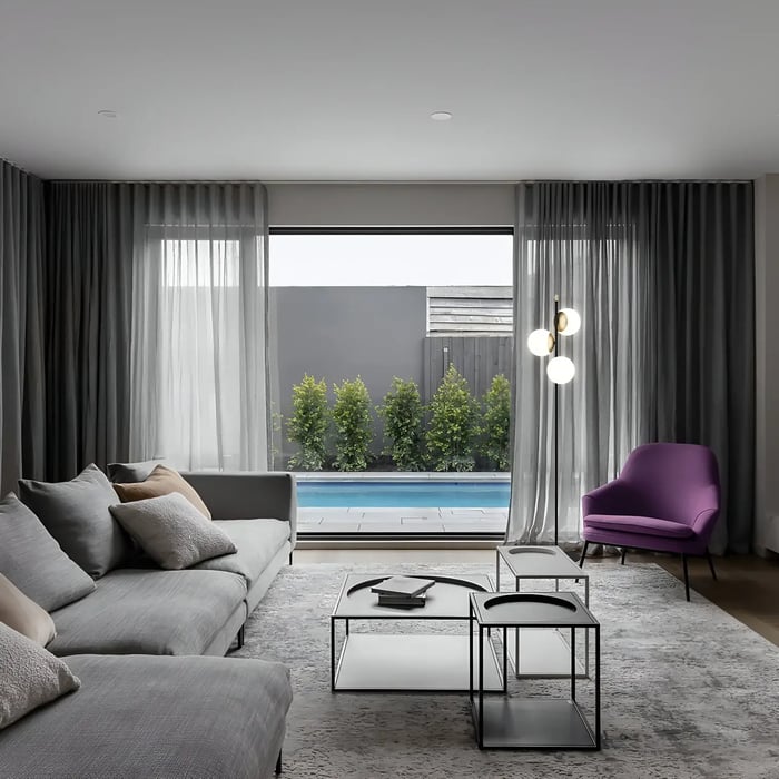 A contemporary living room with a large gray sofa, a purple accent chair, and a view of a pool and greenery through large windows. A multi-globe floor lamp is positioned near sheer grey curtains.