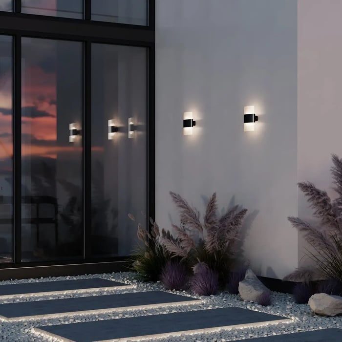 Evening view of a modern patio with black and white wall sconces glowing against a glass façade and decorative gravel garden.