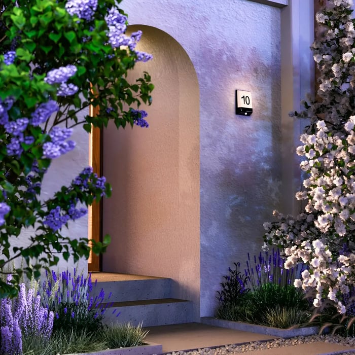 Front door entrance lit by a square wall light with number 10, framed by blooming trees and purple garden flowers.