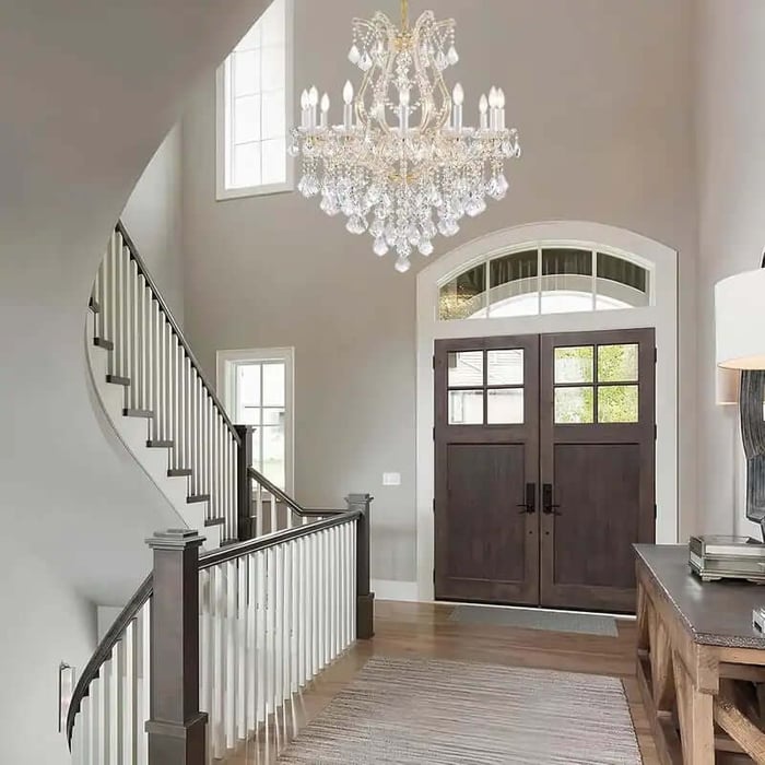 Bright entryway with a large crystal chandelier hanging above a curved staircase, double wooden front doors, and a wooden console table with a lamp.
