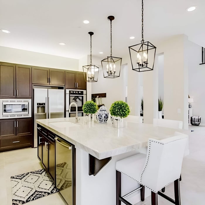Bright modern kitchen with a large marble island, white nailhead barstools, dark wood cabinets, stainless steel appliances, and three black geometric lantern pendant lights hanging above.