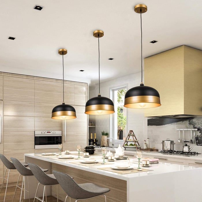 Modern kitchen with a large white island, four gray barstools, and three black-and-gold dome pendant lights hanging above, surrounded by light wood cabinets and a gold range hood.
