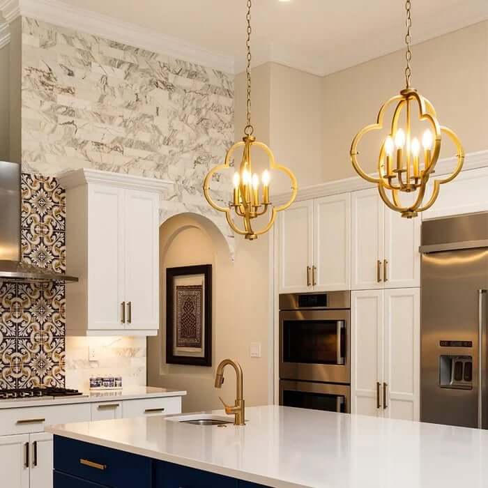 Elegant kitchen with a large white island, blue cabinetry, stainless steel appliances, and two gold quatrefoil pendant lights hanging above, framed by white cabinets and marble and patterned tile backsplashes.