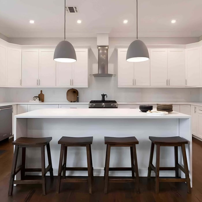 Bright modern kitchen with an all-white island and cabinets, four dark wooden stools, and two large gray dome pendant lights hanging above a central cooktop and range hood.