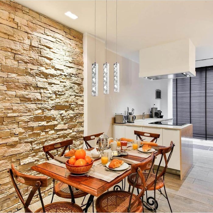 Modern kitchen and dining area featuring a rustic stone accent wall, wooden table with breakfast setup, and three contemporary pendant lights hanging above.