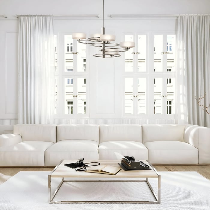  A bright living room with a minimalist aesthetic, featuring a silver multi-tiered chandelier, a large white sectional sofa, and floor-to-ceiling windows with sheer white curtains.