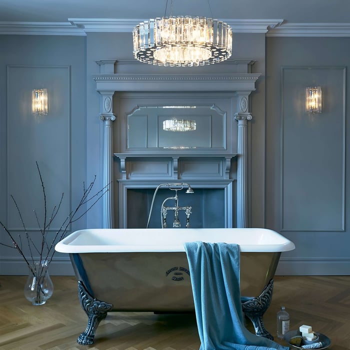 n opulent bathroom featuring a clawfoot tub set against a sophisticated blue-gray backdrop with ornate detailing. An elegant crystal chandelier and matching wall sconces provide a luxurious glow, elevating the classic decor.