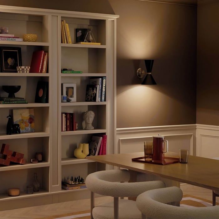 Black hourglass-shaped wall sconce illuminating a warm-toned dining room with open shelving.