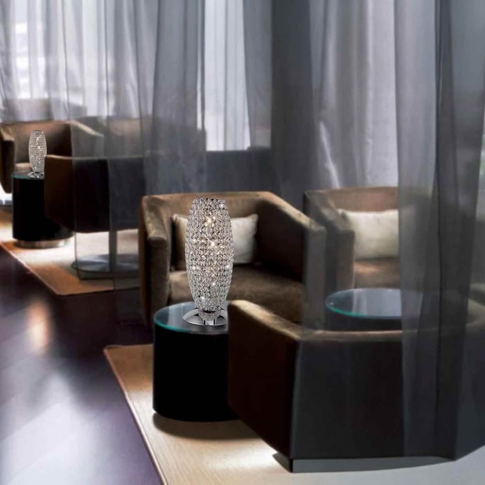 Crystal mosaic table lamp on a black side table between lounge chairs with sheer curtains in the background.