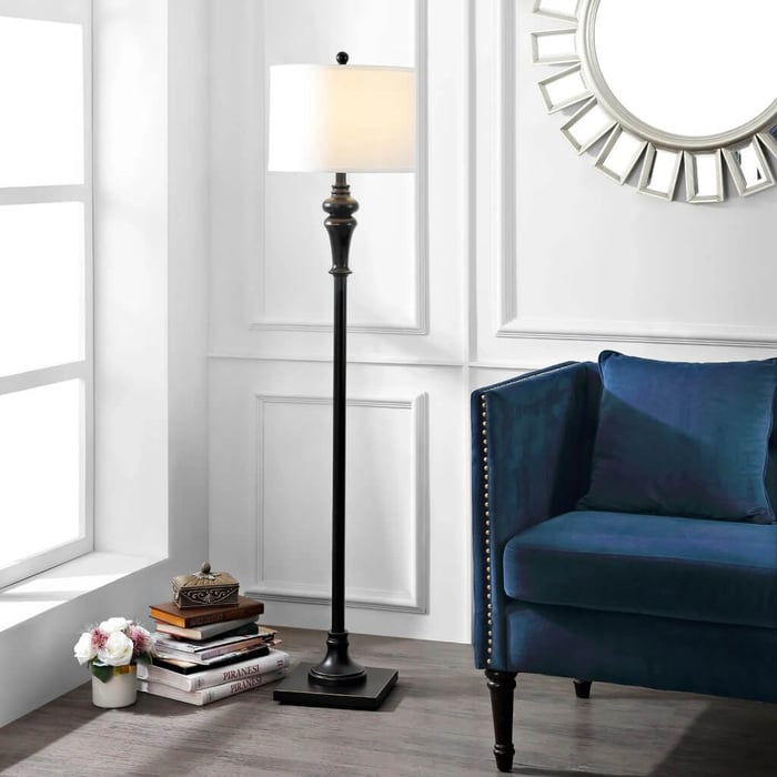 Black traditional floor lamp with white drum shade beside a blue velvet sofa in a white paneled living room with books and flowers.