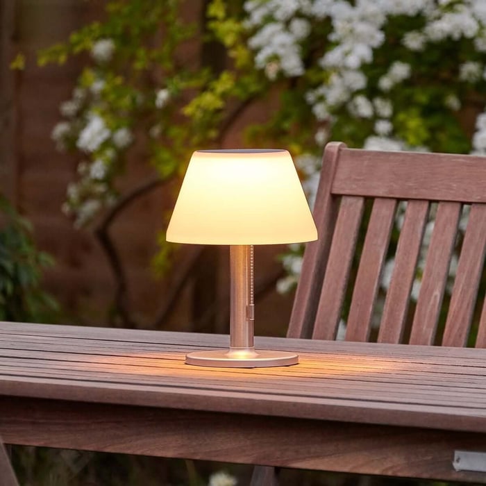 Warm-glow outdoor table lamp on a wooden garden table beside a slatted chair, with flowering plants in the background.