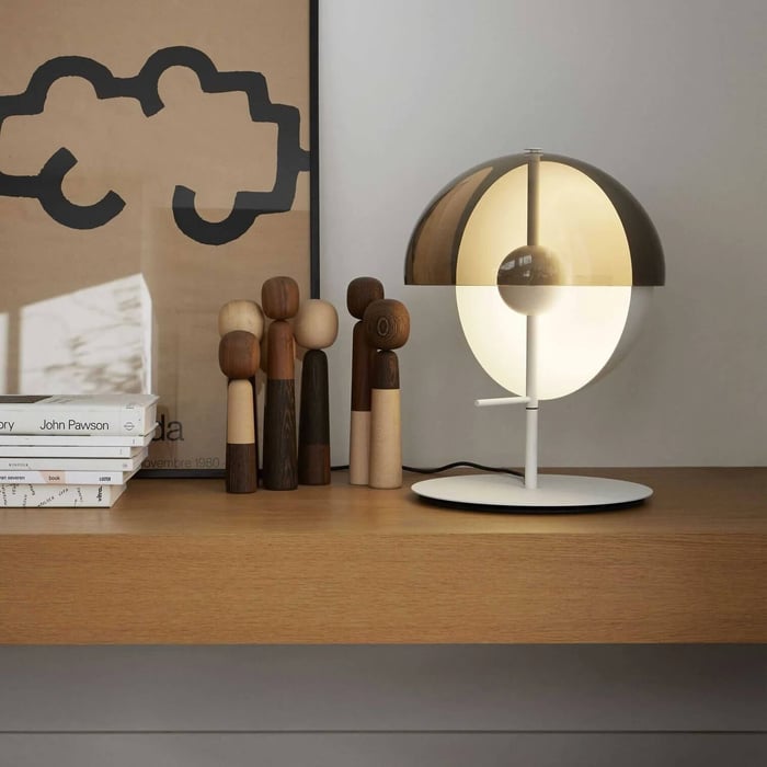 A modern glass orb table lamp with a gold and clear gradient sitting next to abstract wooden peg figures on a light wood shelf.