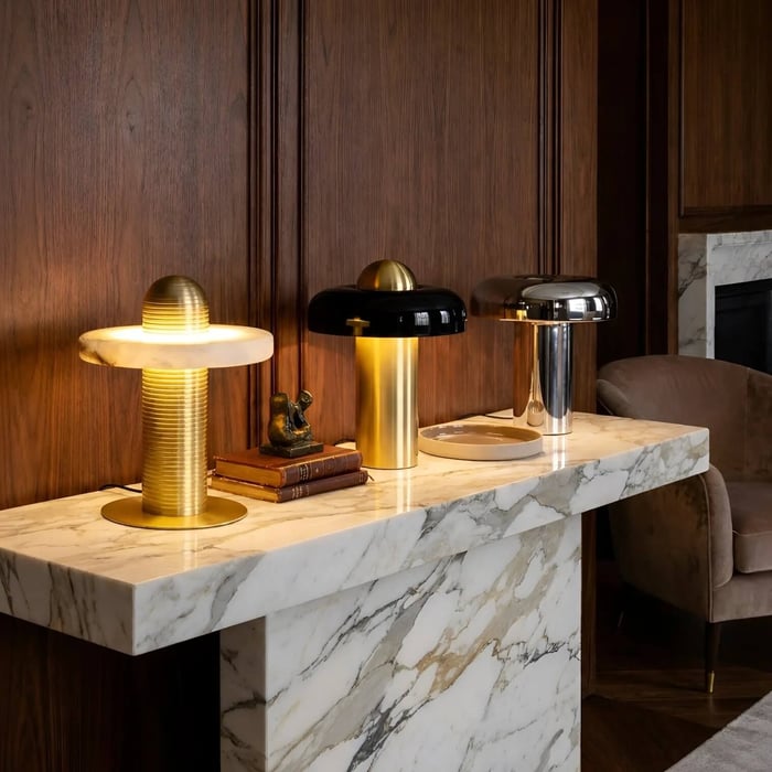 Wood-paneled interior with three sculptural table lamps in gold, black, and chrome displayed on a marble console.