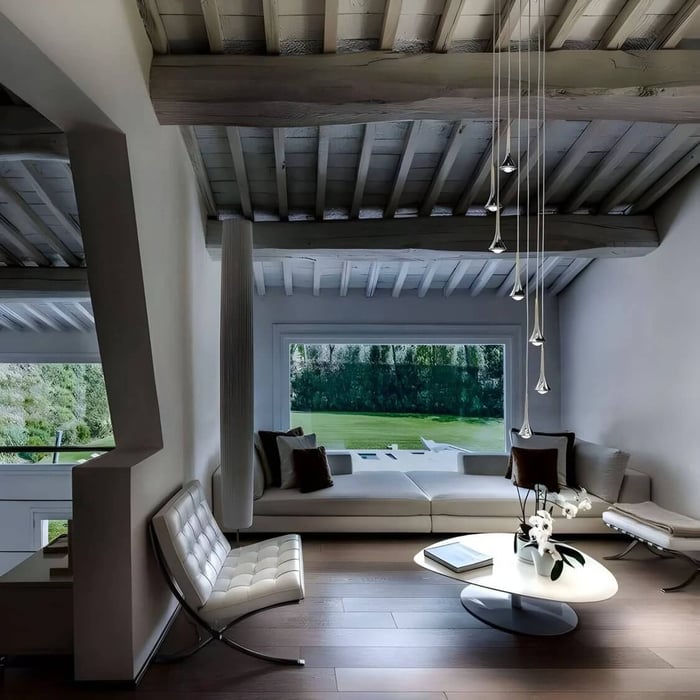 Minimalist living room with exposed wooden ceiling beams, a low white sectional, white lounge chair, round coffee table, and slim multi-drop pendant lights near a large picture window.
