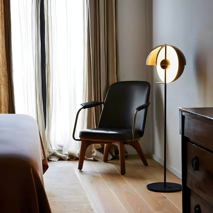Glowing halo-style floor lamp beside a black chair in a calm bedroom with sheer curtains.