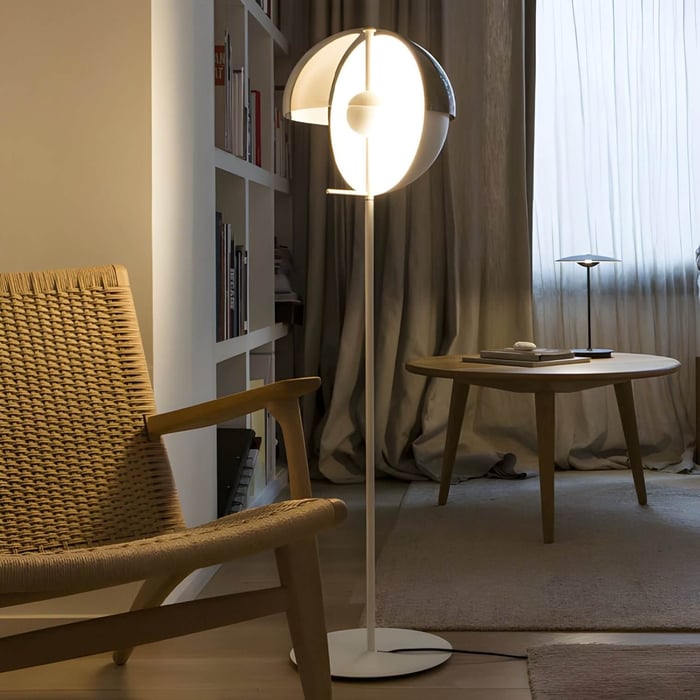 Cozy reading nook with a sculptural floor lamp, wicker lounge chair, wooden side table, and soft curtain-draped window.