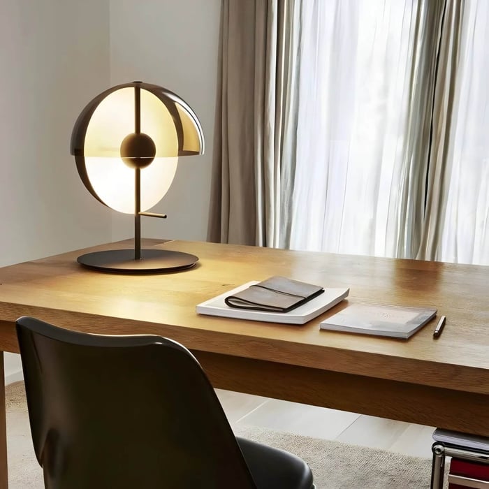 Modern black desk lamp with a circular shade glowing on a wooden desk near sheer curtains and a chair.