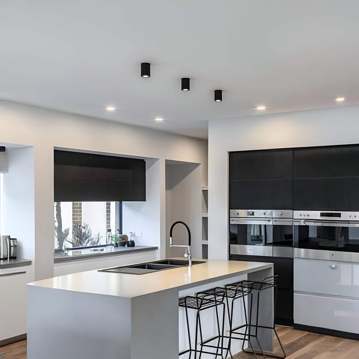 Contemporary white kitchen with a central island, black cylinder surface downlights, and recessed ceiling spots providing bright task lighting.