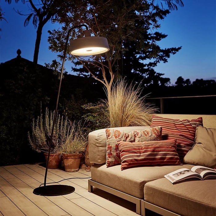 Black outdoor floor lamp with wide shade lighting a patio sofa and potted plants at dusk.