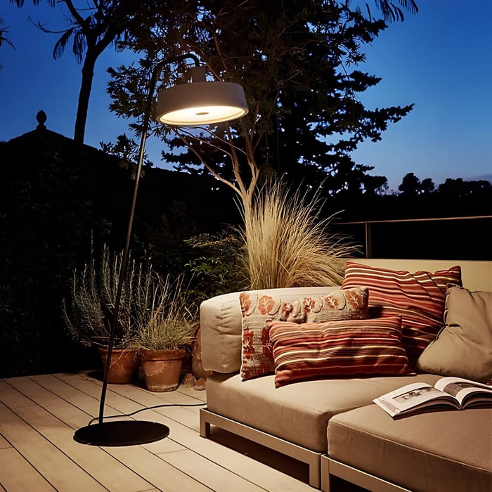 Outdoor floor lamp with wide circular shade beside a beige sofa and striped cushions on a wooden patio at dusk.