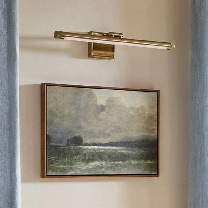 Brass wall-mounted picture light illuminating a framed landscape painting on a pale wall between soft blue curtains.