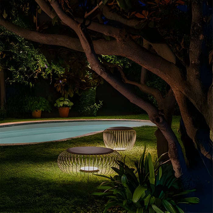 Two low, circular wire-frame outdoor lights with flat tops glowing near a pool and under a large tree.