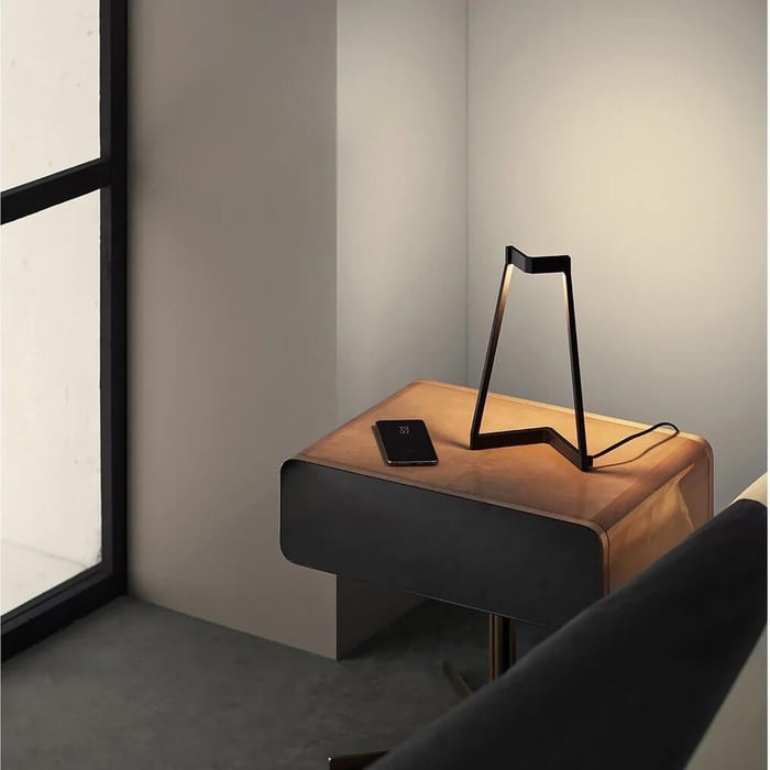 Minimal black geometric table lamp on a wooden bedside table in a softly lit modern room.