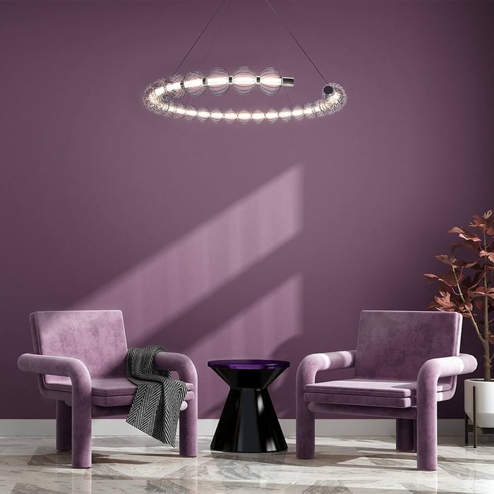 Circular bubble chandelier glowing against a purple wall with two velvet chairs and a black side table.