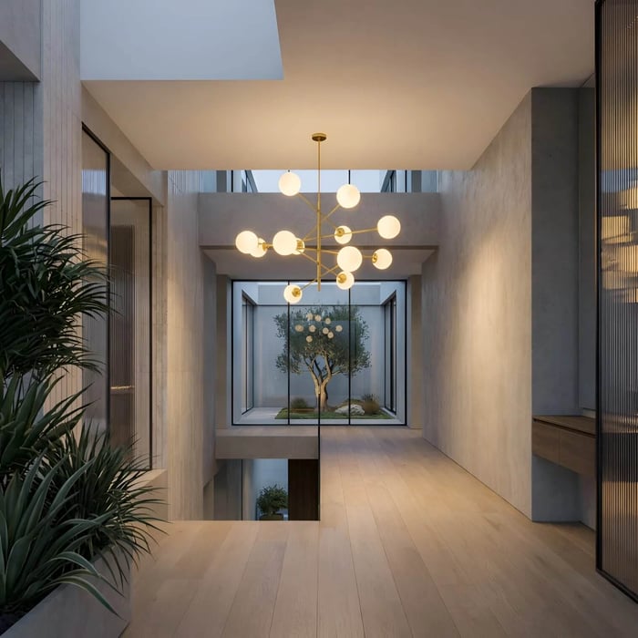 A modern architectural hallway with light wood floors and grey concrete walls. An oversized, spiky gold chandelier with white globe lights serves as a focal point, with a glass-walled courtyard containing an olive tree visible in the background.
