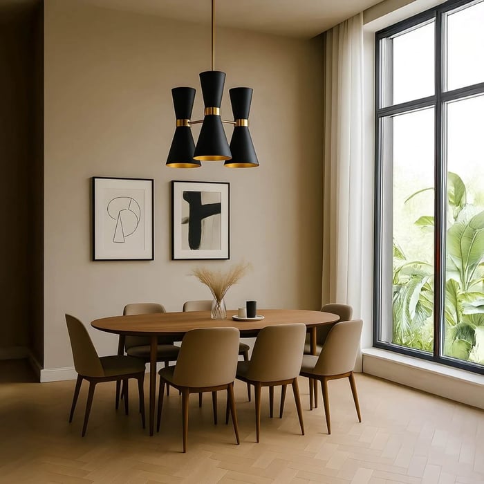 A warm, neutral dining room featuring an oval wooden table and six beige upholstered chairs. A striking matte black chandelier with three hourglass-shaped shades and gold interiors hangs above the table, flanked by two pieces of abstract framed art.