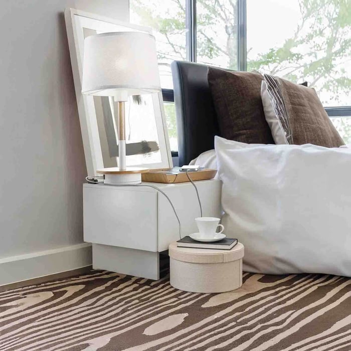 Minimal white table lamp on a wall-mounted bedside table next to a bed, with a tray and charging cable.