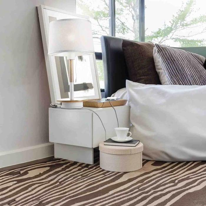 A modern bedside table with a white lamp sits next to a neatly made bed by a large window. A small round ottoman with a cup and book rests on a patterned rug.