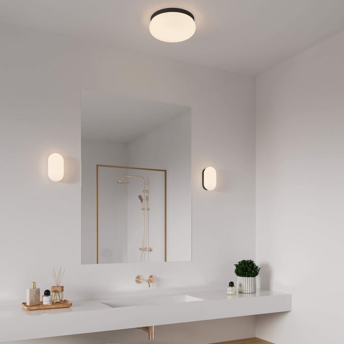 Simple white bathroom with a single mirror, modern black faucets, round ceiling light, and two capsule-shaped wall lights.