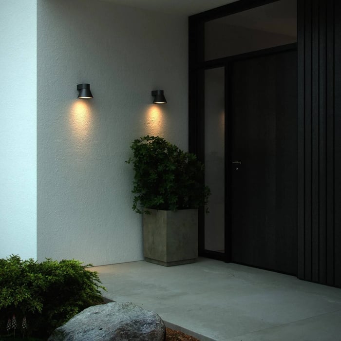 Two compact outdoor wall lights casting warm downward beams beside a modern front door and planter.